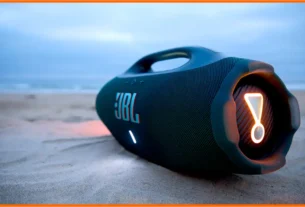 Alto-falante portátil JBL em uma praia de areia, com o mar ao fundo e luz suave ao entardecer.